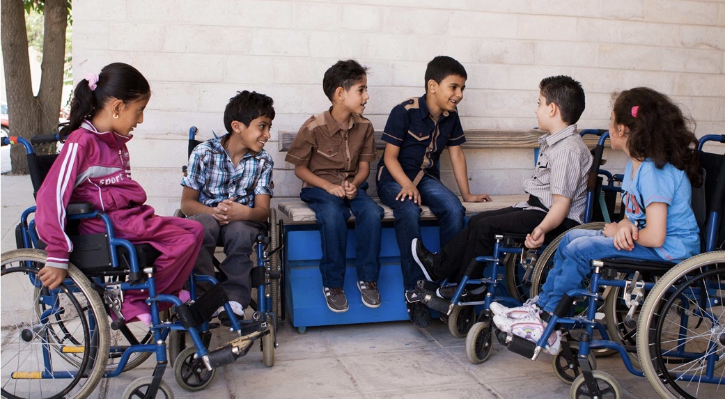A group of children sit in a semi-circle talking and smiling. Four of the children are sitting in wheelchairs.