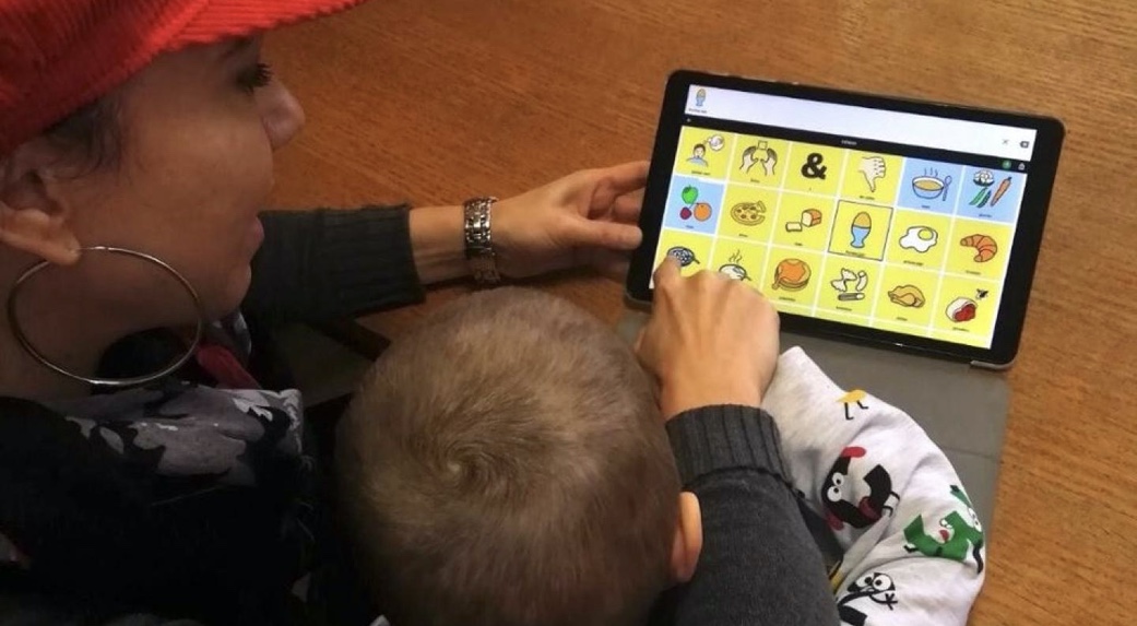 A woman and young boy are using a tablet with the cboard application. It depicts a grid of colored tiles and symbols.