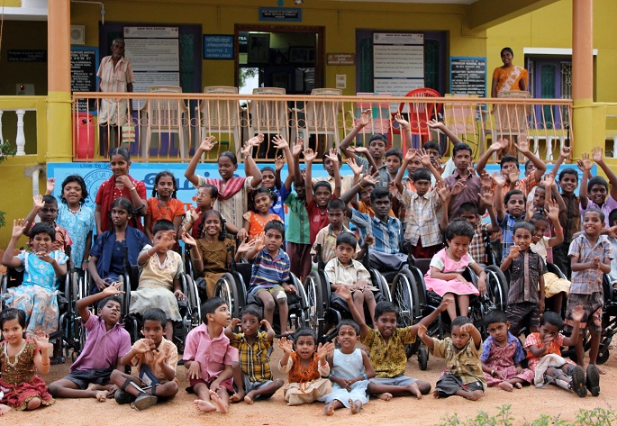 A large group of children in a courtyard waving. Many of the children are in wheelchairs